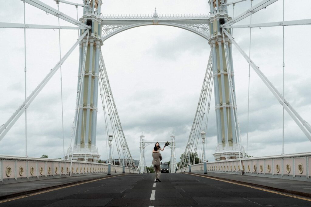 Groom lifting bride on Chelsea Bridge in London, wide shot with dramatic sky, captured by a London wedding photographer.
