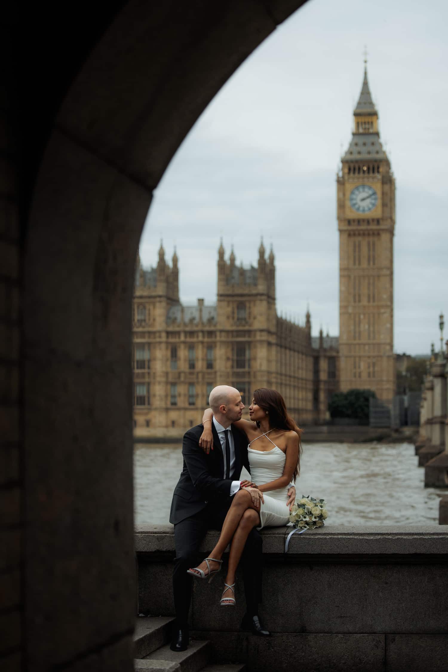 Couple sitting by the Thames with Big Ben framed by an arch during a pre-wedding photoshoot in London.