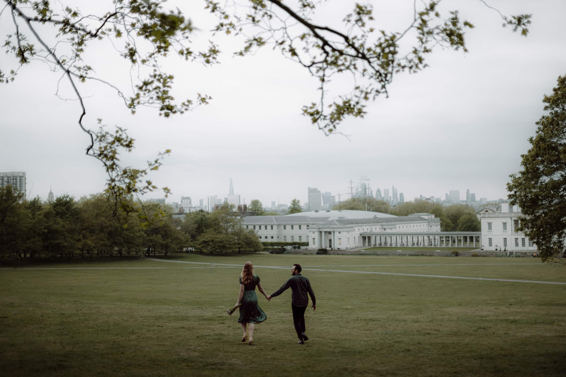 Couple walking hand in hand across Greenwich Park with London skyline during a pre-wedding photoshoot in London.