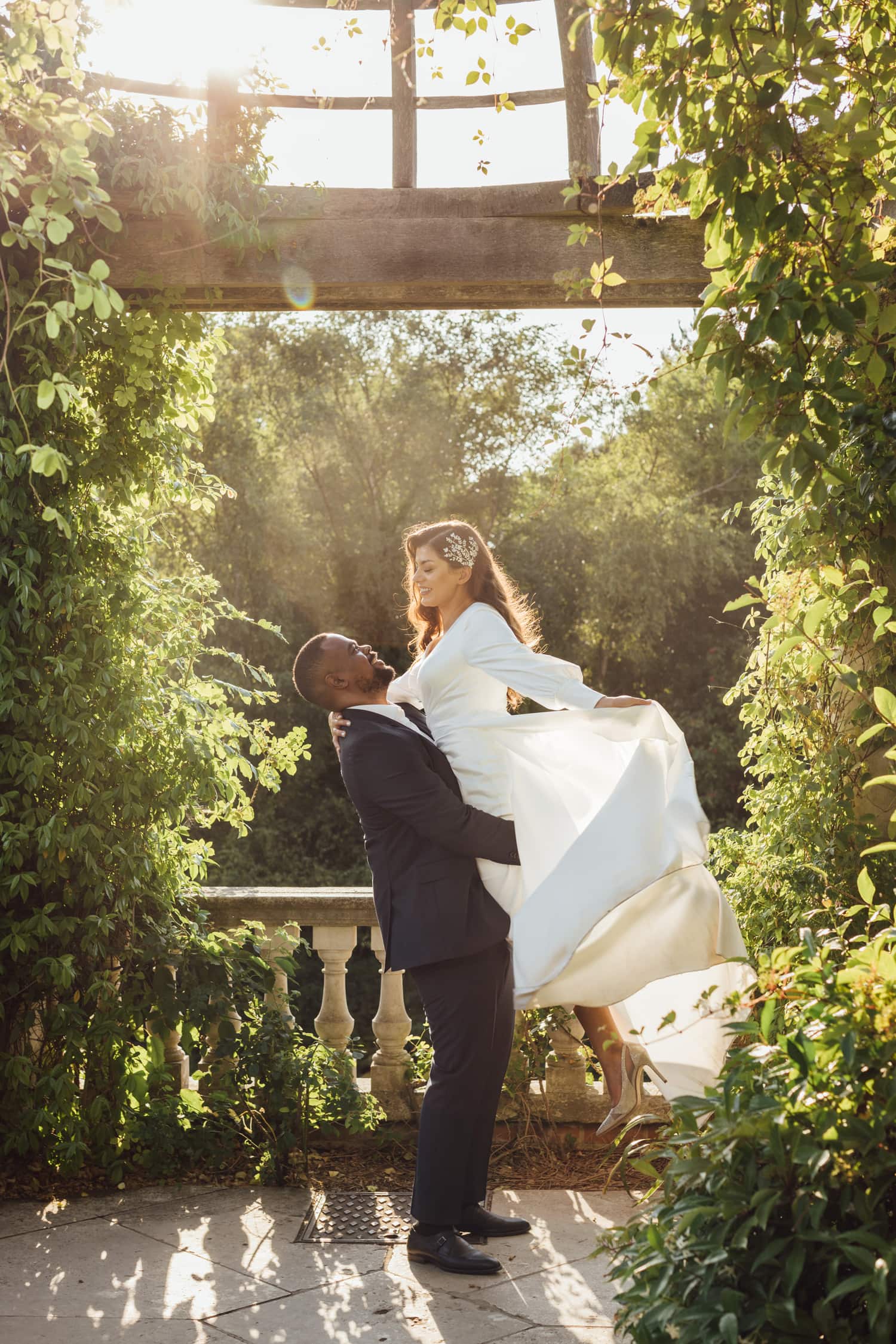 Groom lifting bride under pergola with warm sunlight at Pergola and Hills Garden during a pre-wedding photoshoot in London.